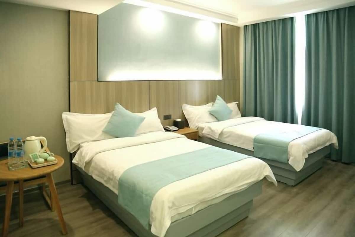 A hotel room with two beds, a bedside table with a lamp, a small table with a tray of items, and a large window with curtains.