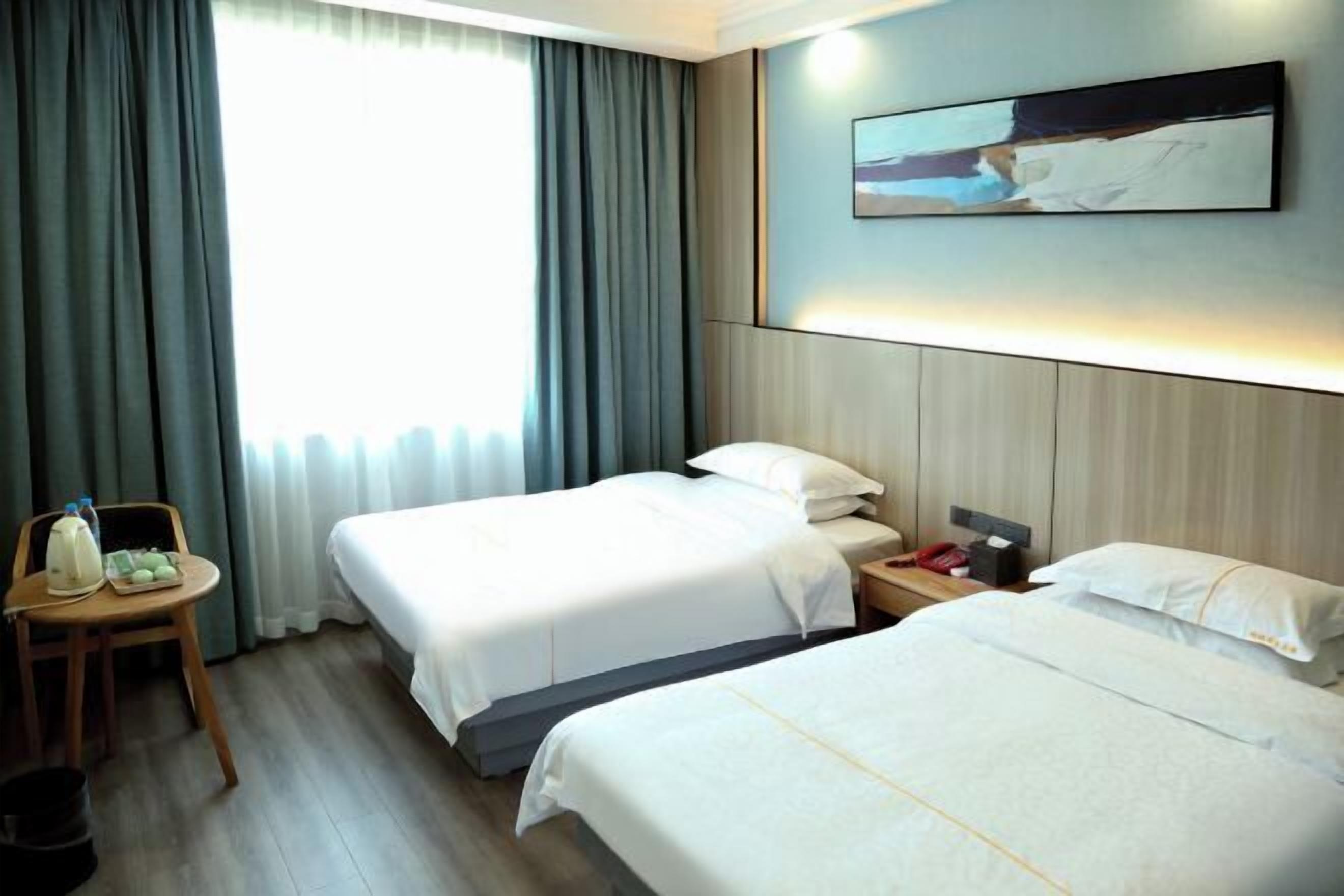 A hotel room with two beds, a small table with a chair, a bedside table with a lamp, and a painting on the wall.