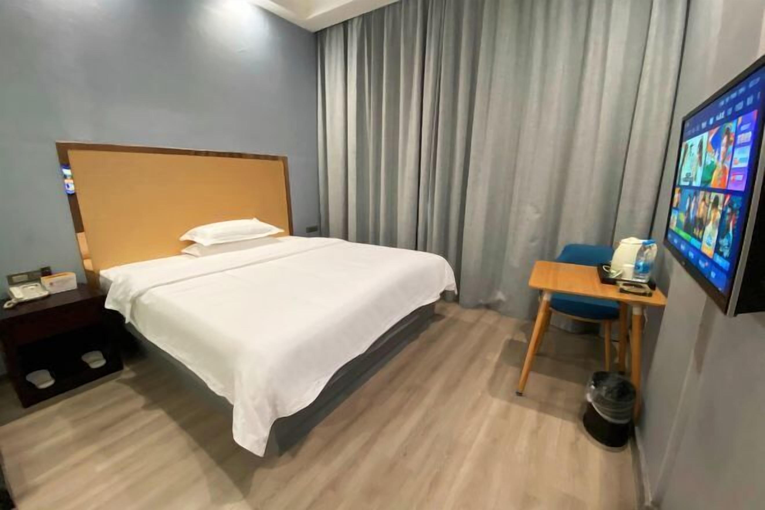 A hotel room with a bed, a desk, a chair, a TV, and a trash bin.
