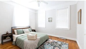 Une chambre à coucher comprenant un lit, des tables de chevet, un ventilateur de plafond, une fenêtre avec des stores, un tableau encadré et un tapis à motifs floraux.