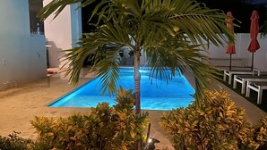 En pool med ett palmträd i mitten, omgiven av ett trädäck och låga buskar.