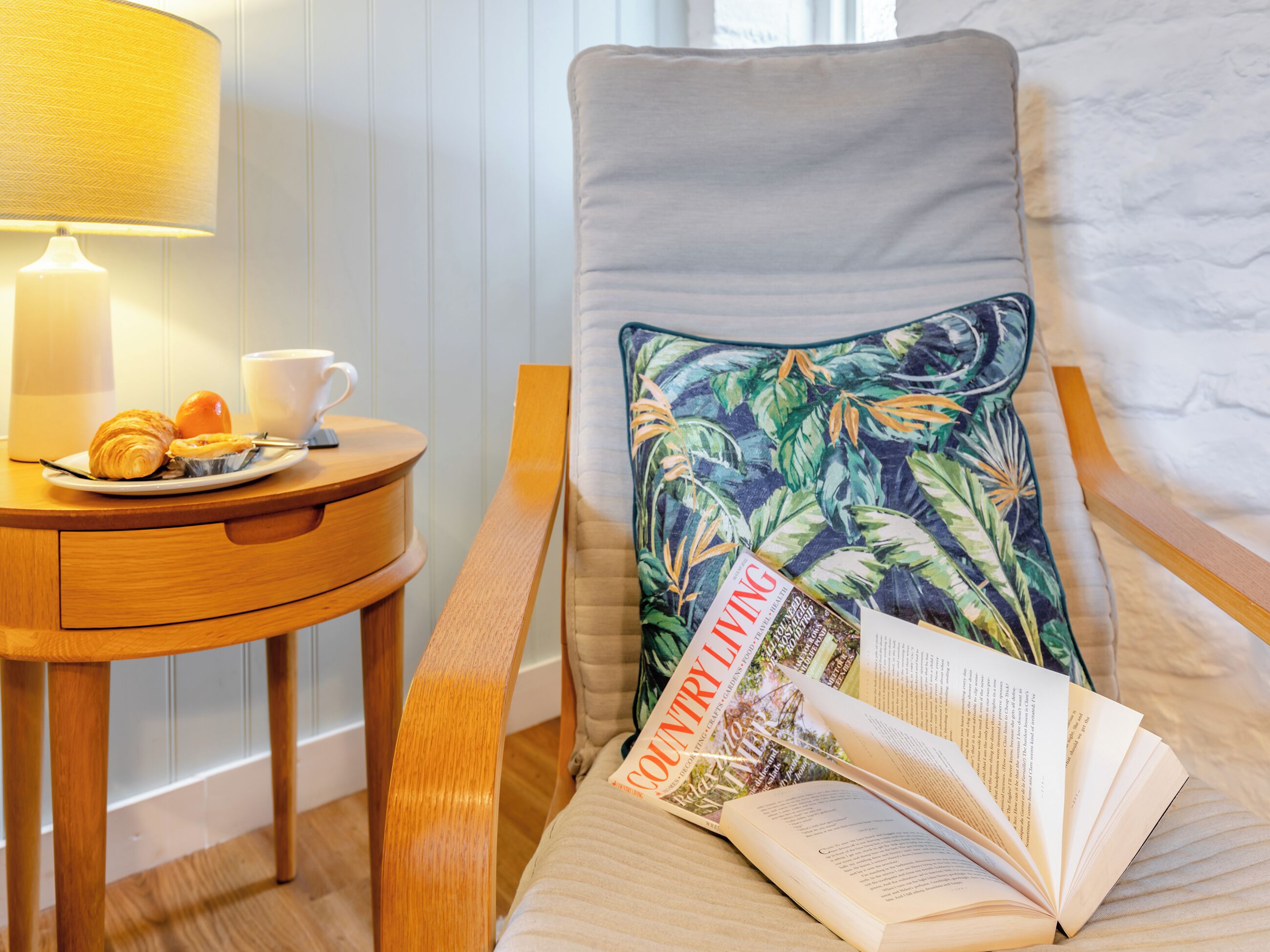 A cozy reading nook with a chair, a table with a lamp, a plate of croissants, a cup, and a book.