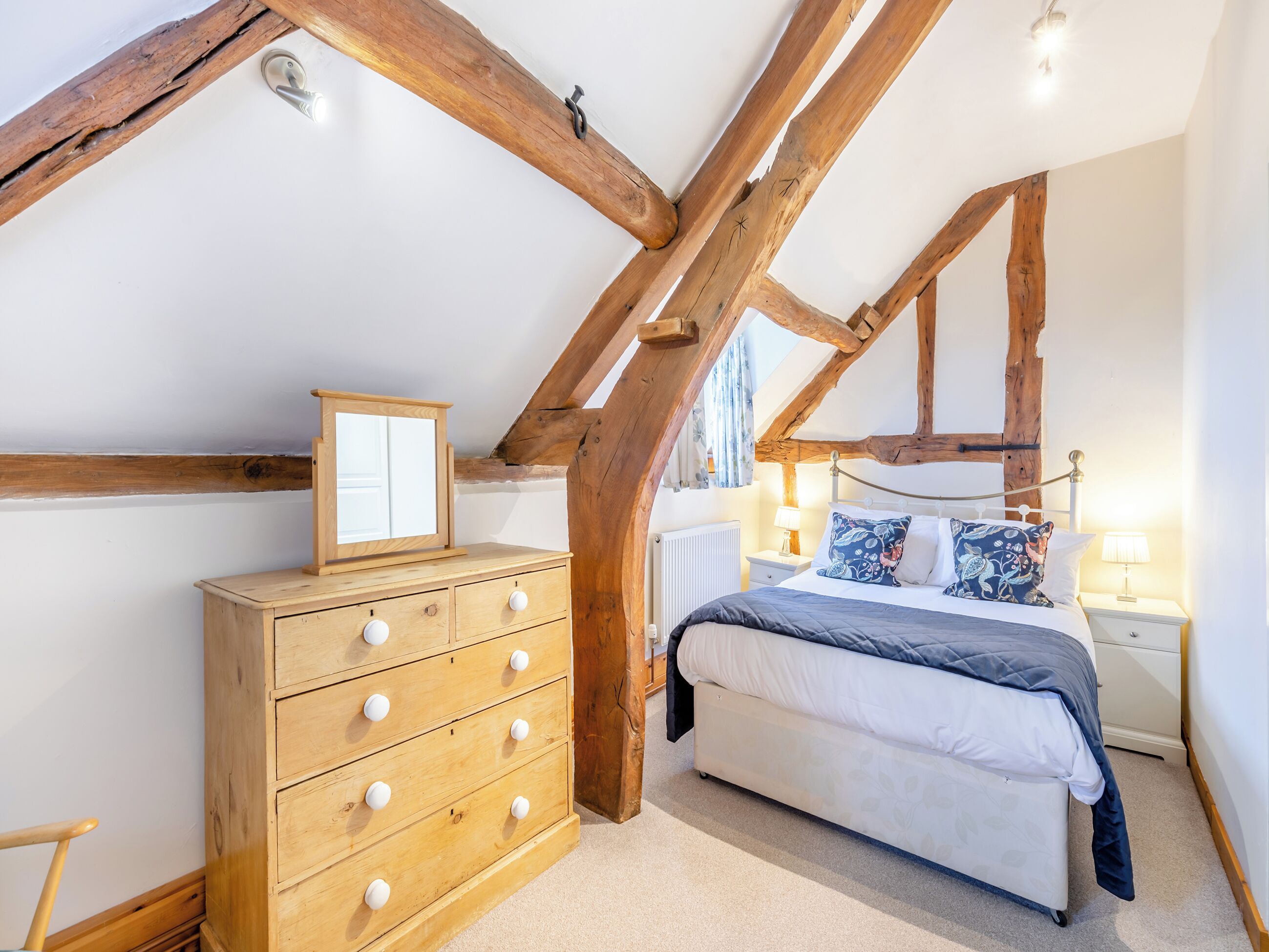A bedroom with exposed wooden beams, a wooden dresser with drawers, a bed with a blue blanket, and a nightstand with a lamp.