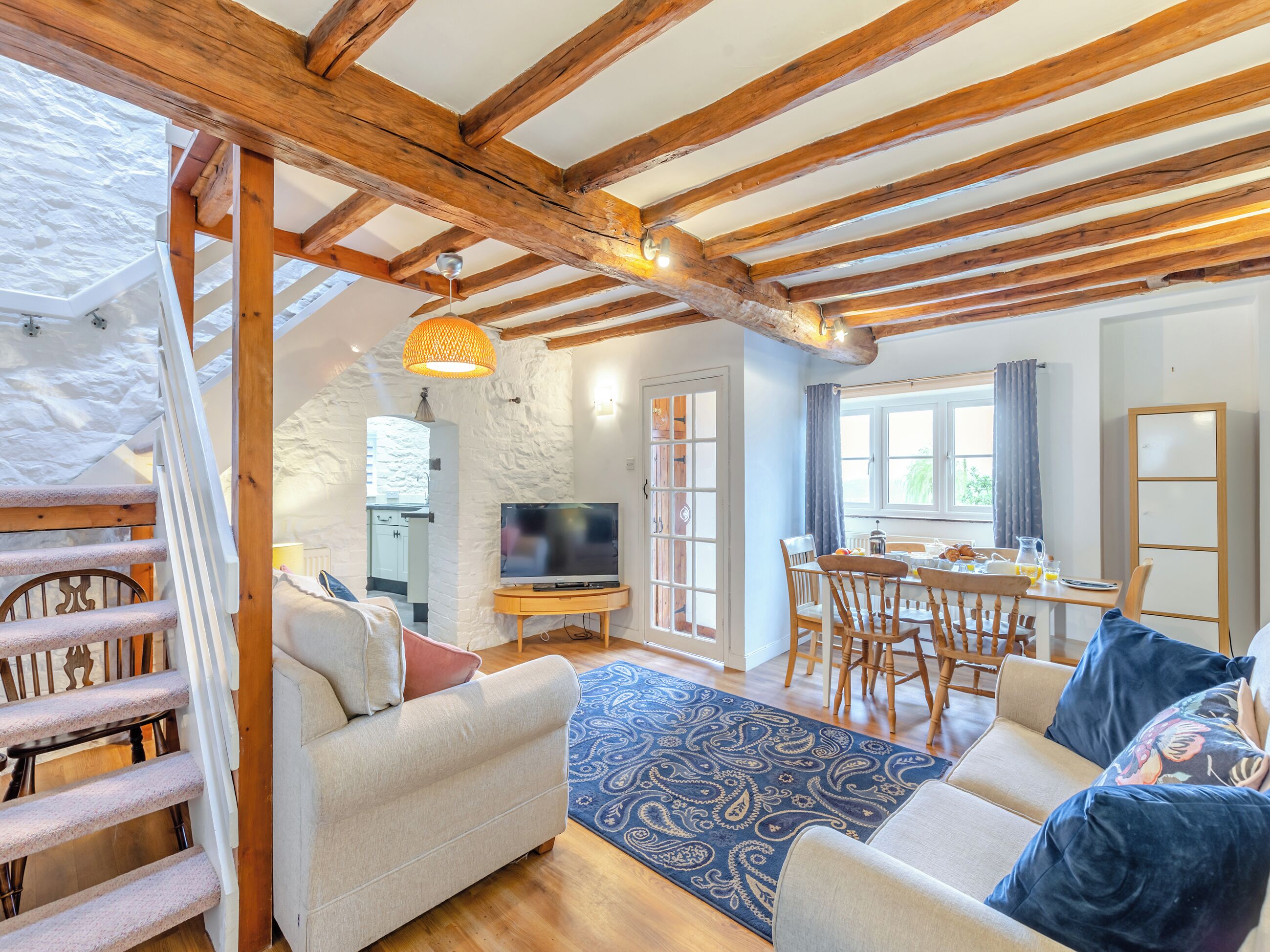 A cozy living room with a wooden ceiling, a TV on a wooden stand, a dining table with chairs, and a staircase.
