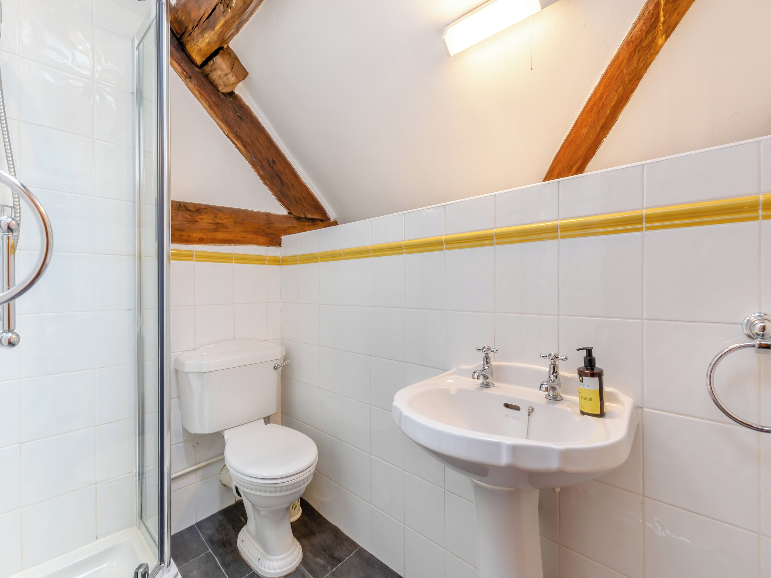 Casa de banho com azulejos brancos, sanita branca, lavatório com dispensador de sabonete amarelo e zona de duche.