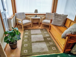 Une pièce chaleureuse avec un plancher en bois, un tapis vert et blanc, deux fauteuils à carreaux, une petite table avec une lampe, un banc en bois et une plante en pot.