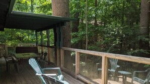 A covered deck with wooden furniture and glass railings overlooking a forest.