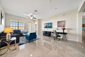 A spacious living room with a ceiling fan, a sofa, a blue armchair, a coffee table, and a desk with a lamp.