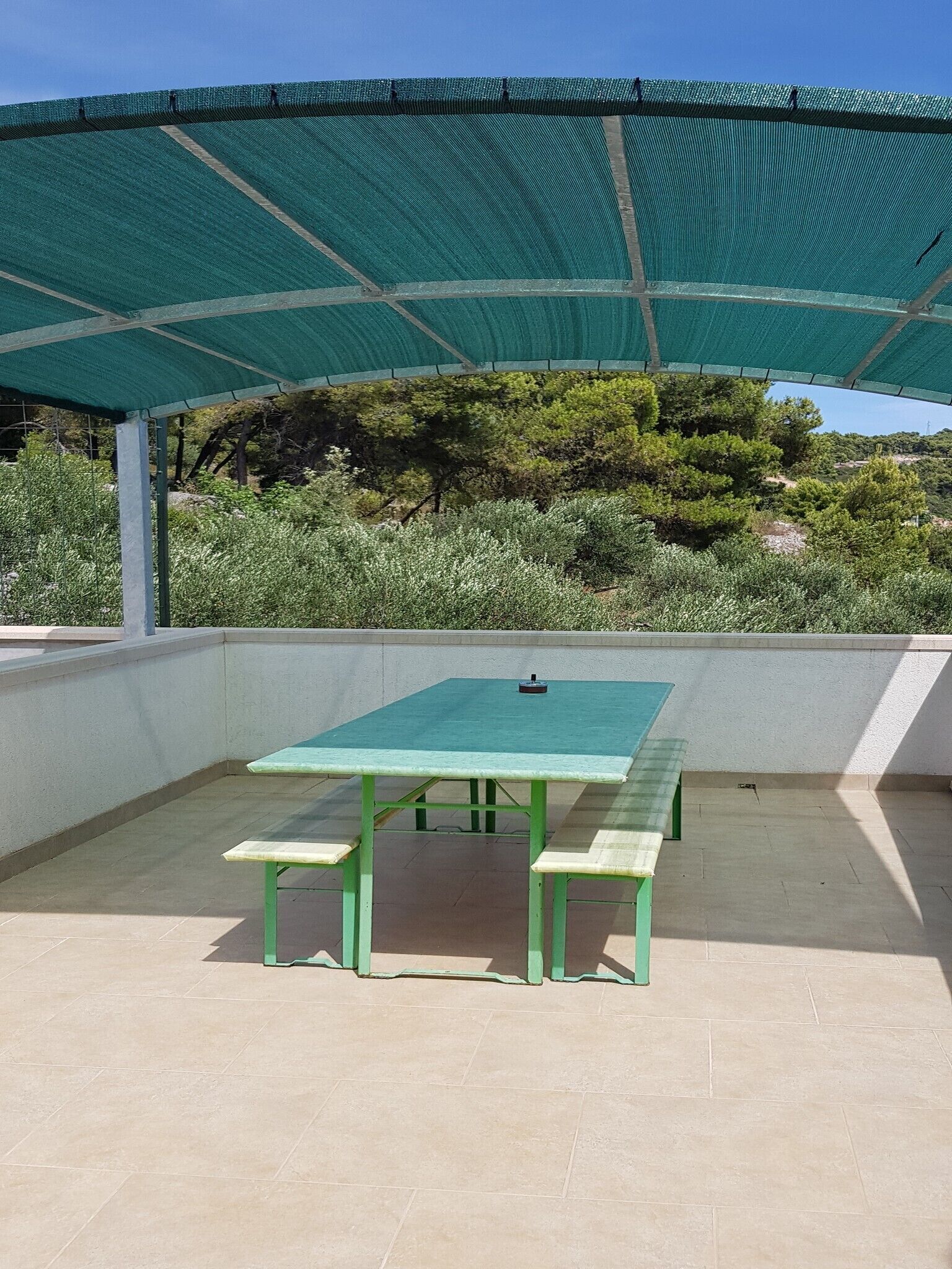 Eine Terrasse im Freien mit einem grünen Tisch und Bänken, einem überdachten Bereich mit einem Netzdach und Bäumen und Sträuchern im Hintergrund.