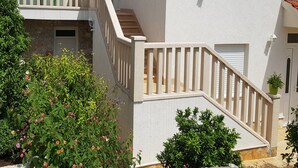 Un edificio de dos pisos con fachada blanca, una escalera de madera con barandilla y un pequeño balcón con sombrilla verde.