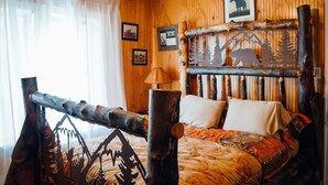 A rustic room with wooden walls, a bed with patterned bedding, a wooden headboard with carved designs, and a window with sheer curtains.