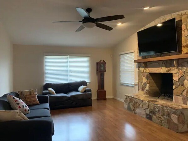 A spacious living room with a stone fireplace, a ceiling fan, and a flat-screen TV mounted above it.