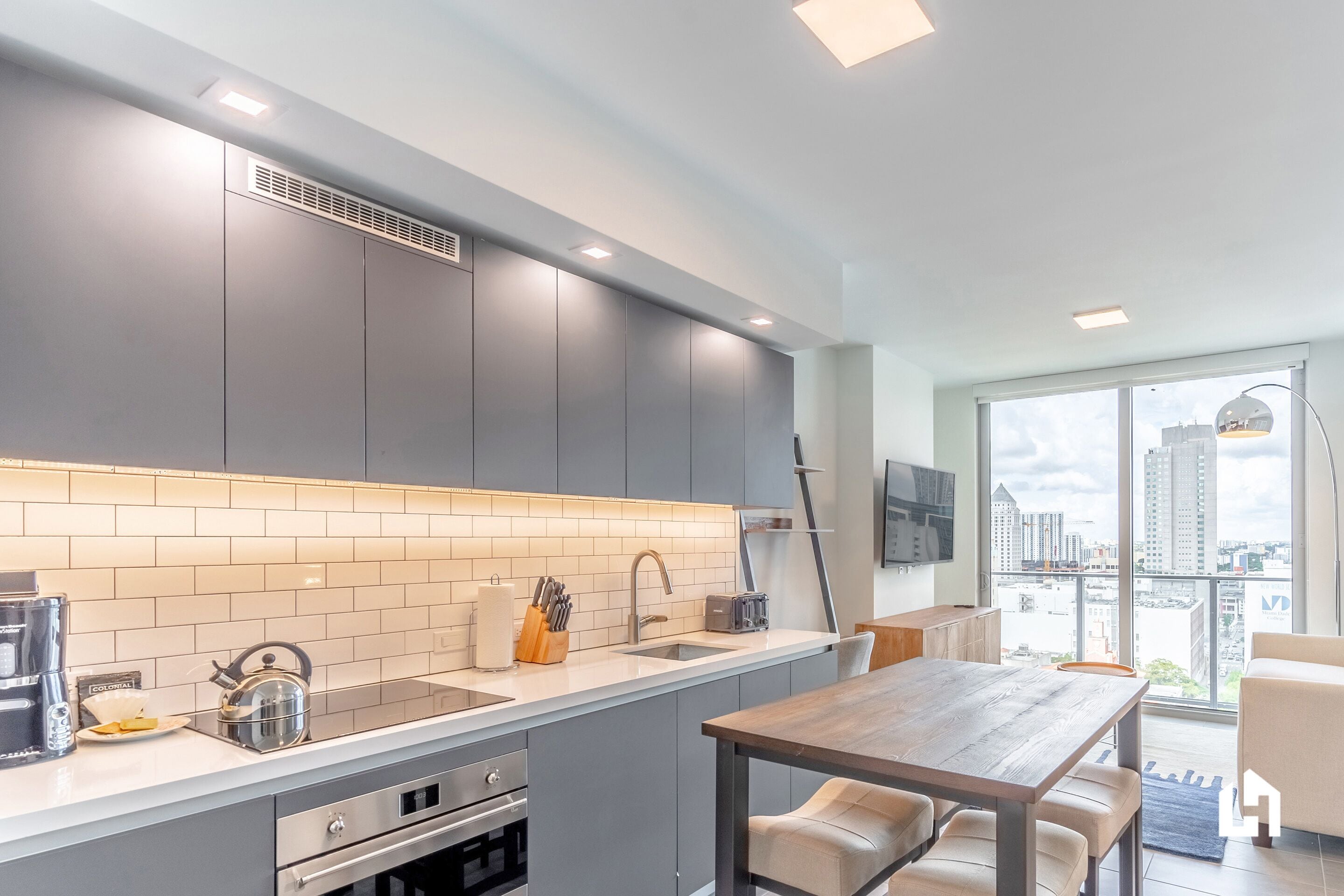 Een moderne keuken met grijze kastjes, een wit aanrechtblad en een groot raam met uitzicht op de stad.