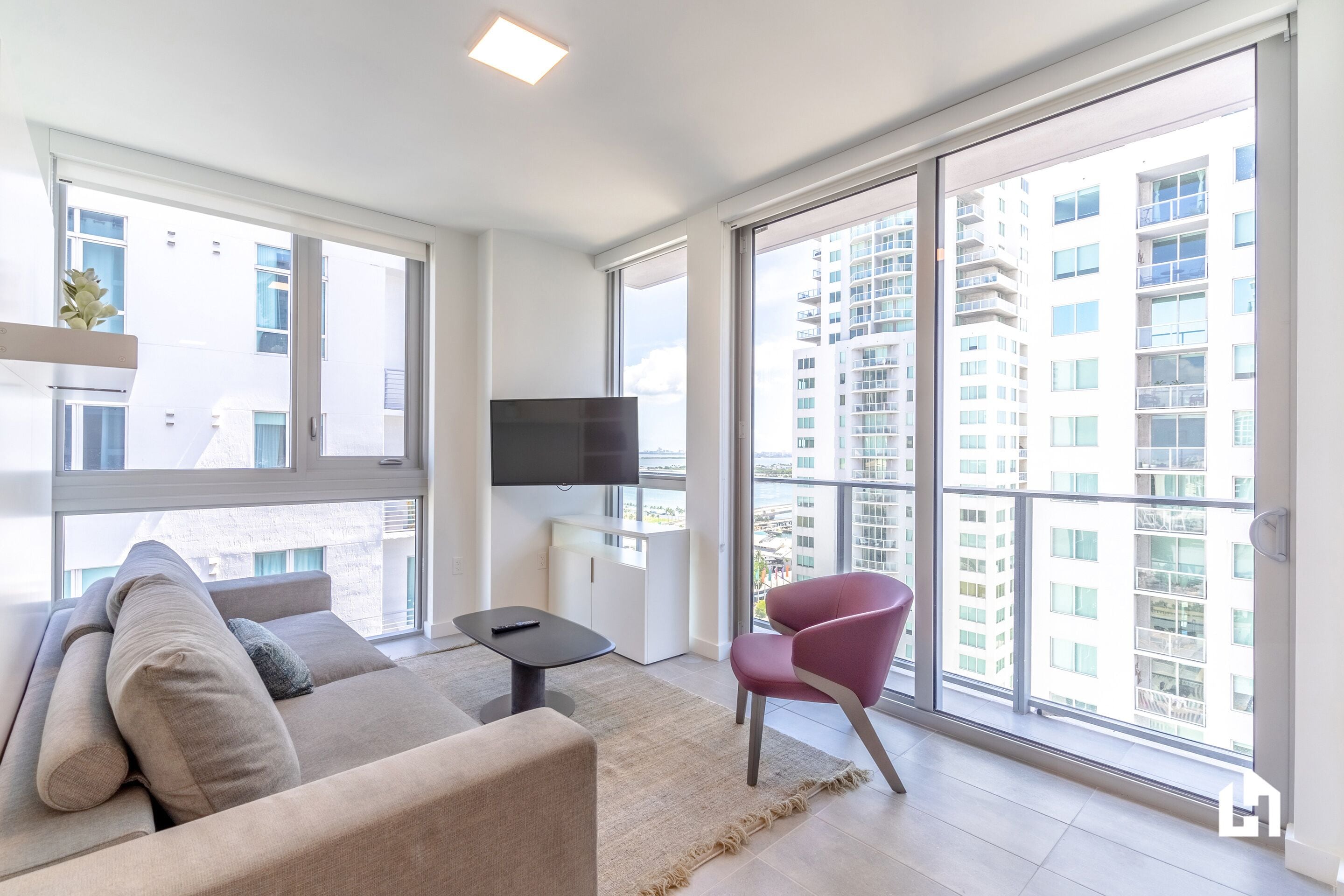 A modern living room with a sofa, a chair, a TV, and large windows offering a city view.