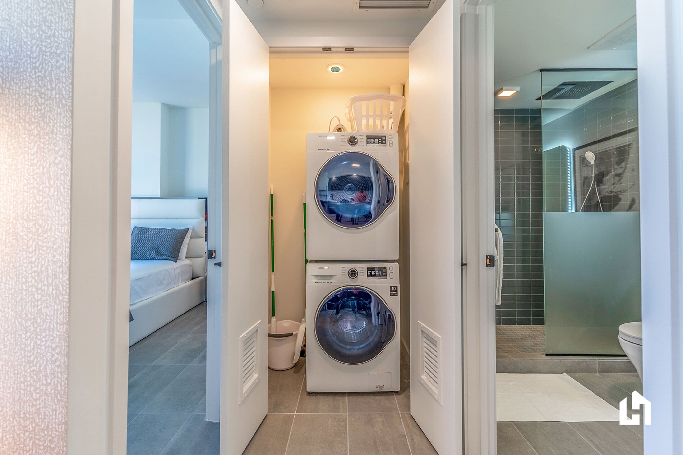 A laundry room with stacked washers and dryers, a bed with pillows, and a bathroom with a glass shower.