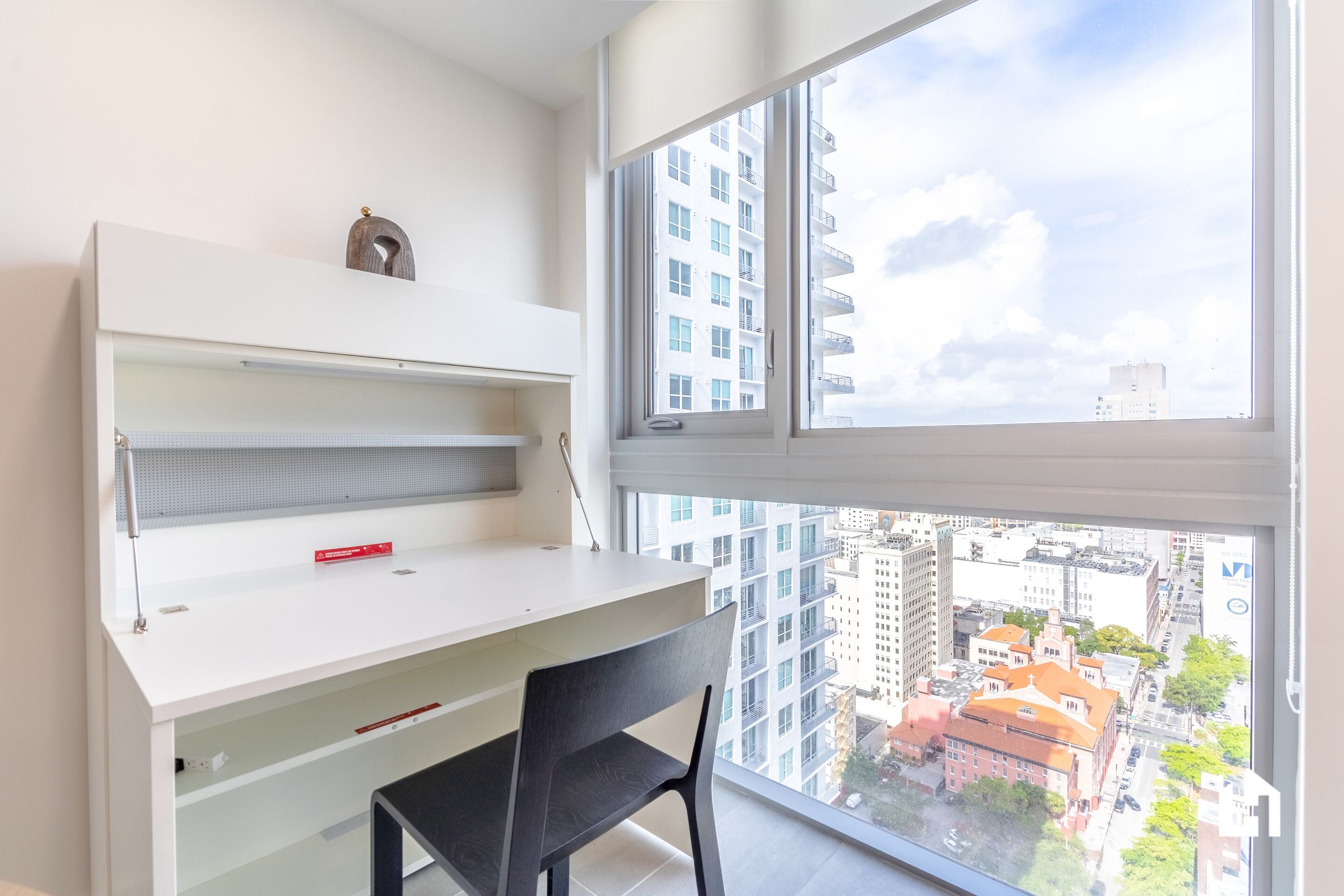 Una habitación moderna con un escritorio blanco, una silla negra y un amplio ventanal con vista a la ciudad.