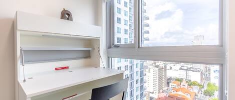 Una habitación moderna con un escritorio blanco, una silla negra y un amplio ventanal con vista a la ciudad.