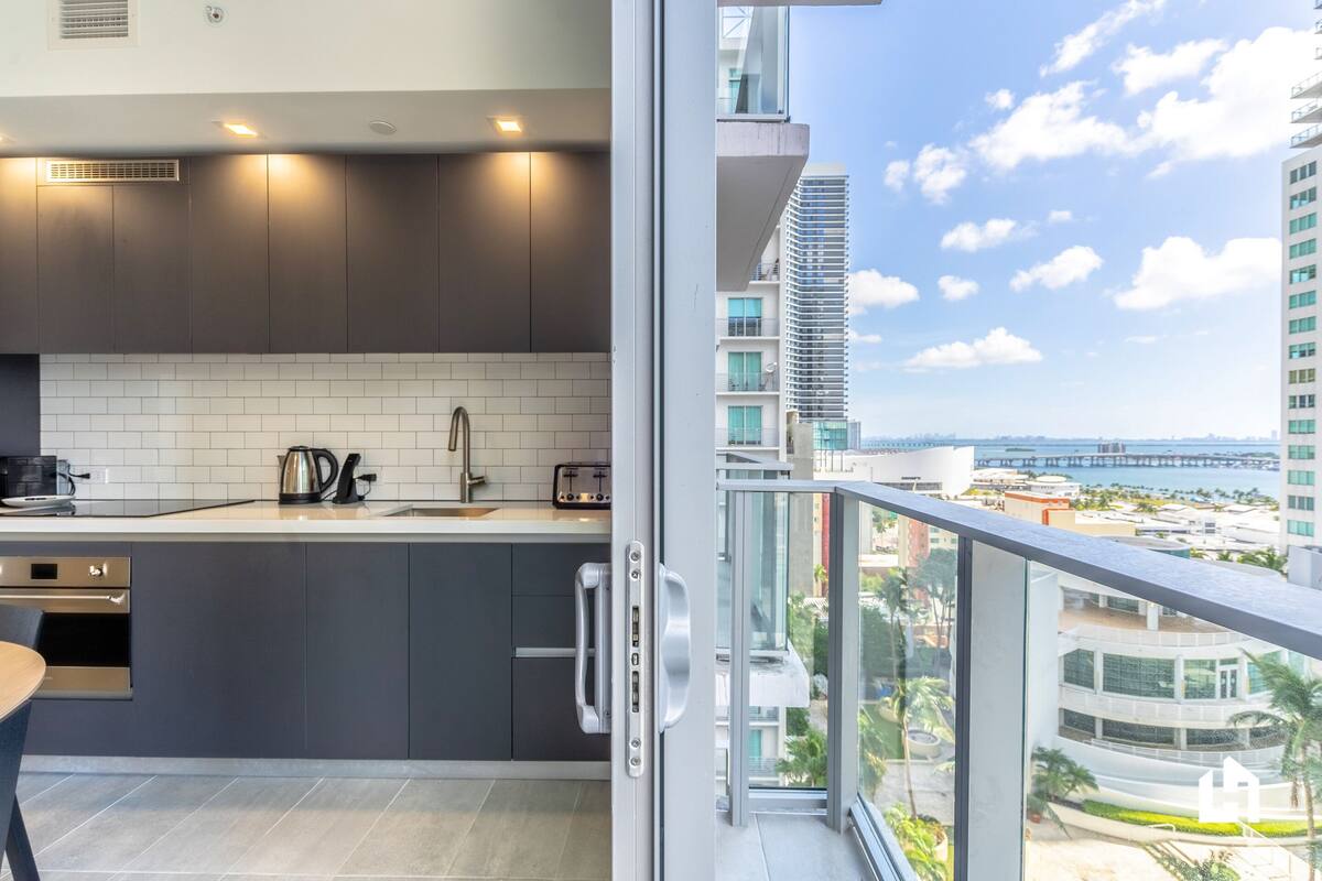Una cocina moderna con armarios oscuros, baldosas blancas y vistas a un paisaje urbano desde un balcón.