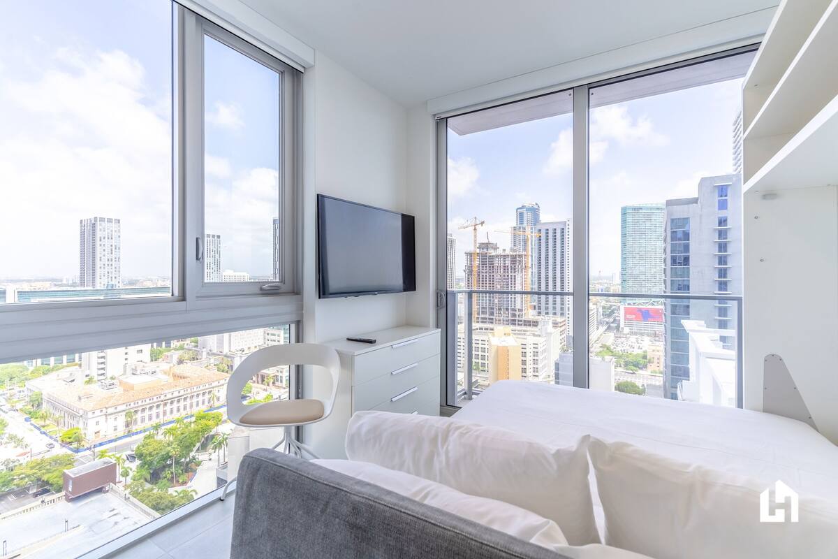 Habitación de hotel moderna con una cama grande, un escritorio con televisor de pantalla plana, una silla y vistas a la ciudad a través de amplias ventanas.