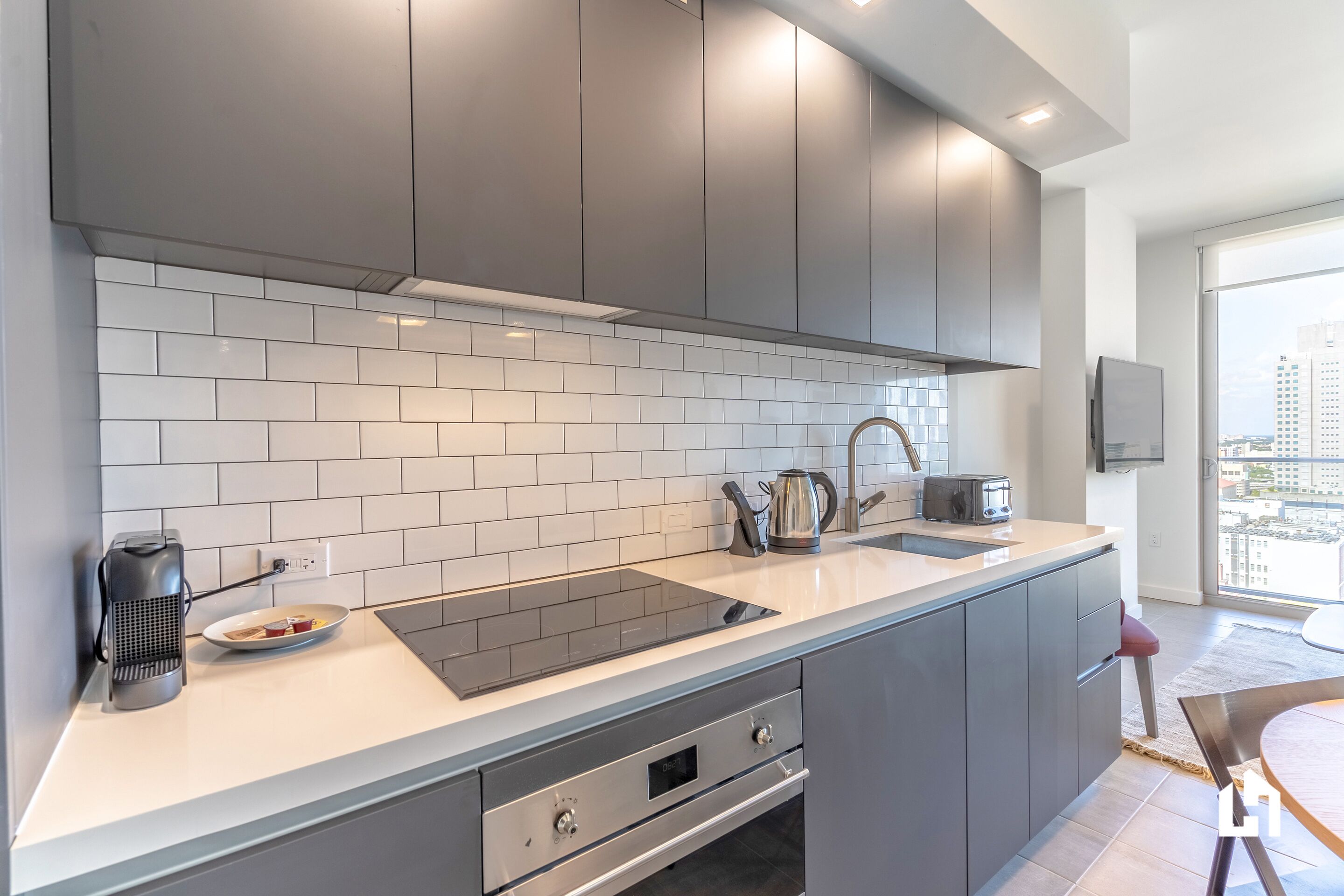 A modern kitchen with grey cabinets, a white tiled backsplash, a built-in stove, and a large window offering a city view.