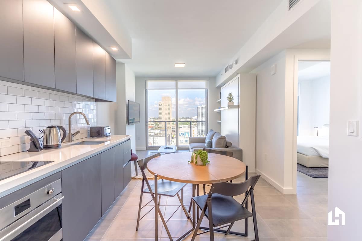Una cocina moderna con armarios grises, una encimera blanca y una mesa redonda de comedor con cuatro sillas. Hay un ventanal que ofrece vistas panorámicas de la ciudad.