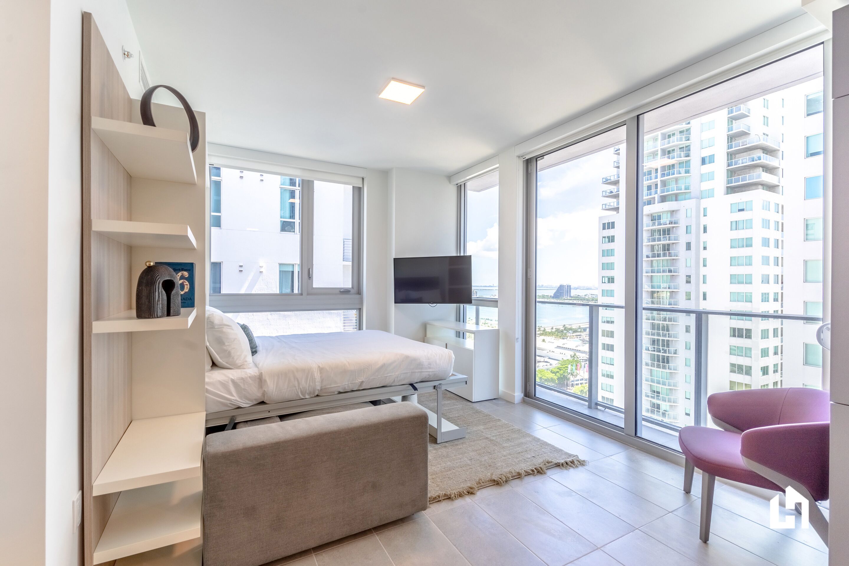 Un dormitorio moderno con cama, televisor, sofá y ventanas amplias con vistas a la ciudad.