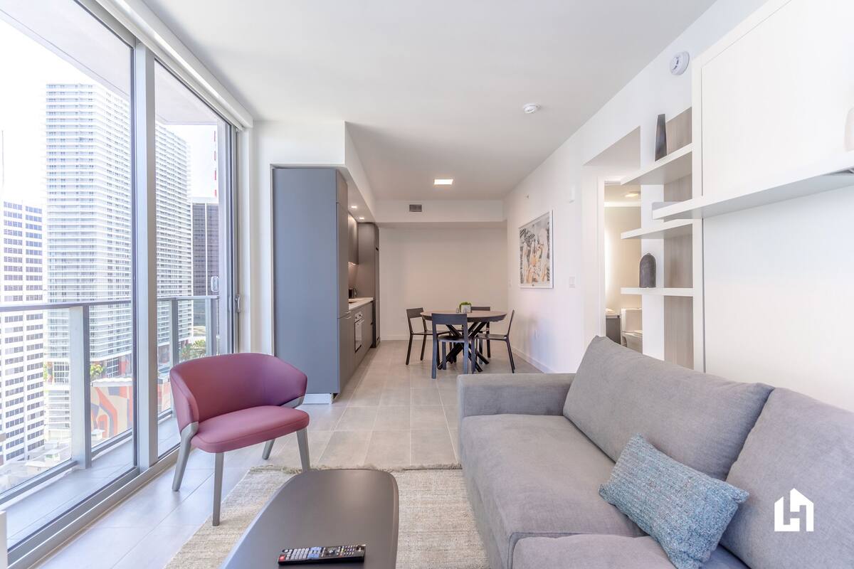 Una sala de estar moderna con un sofá gris, un sillón rojo y una mesa de centro. Se ve una zona de comedor con una mesa y sillas al fondo.