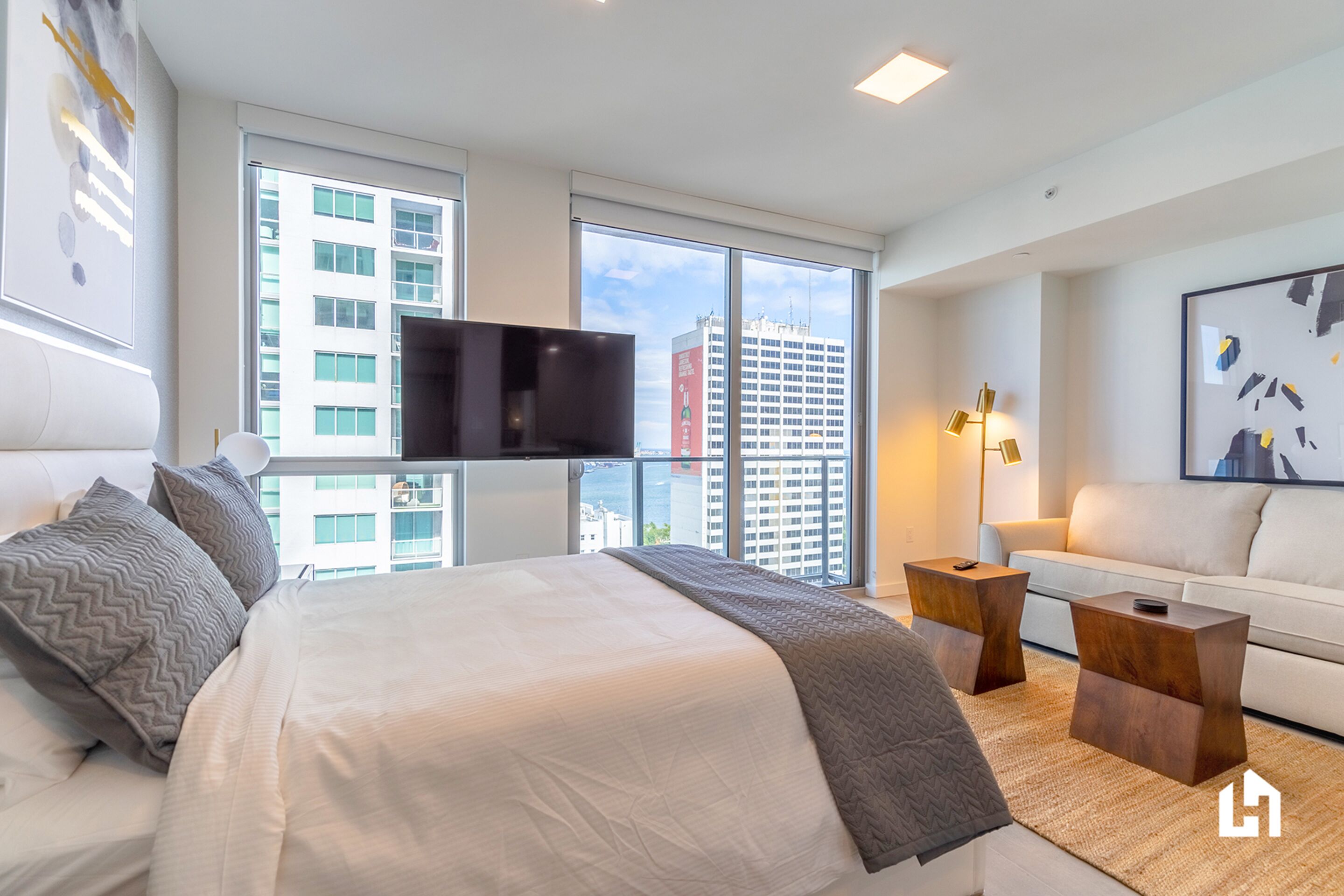 A modern hotel room with a large bed, a flat-screen TV, a sofa, and a view of the cityscape through large windows.