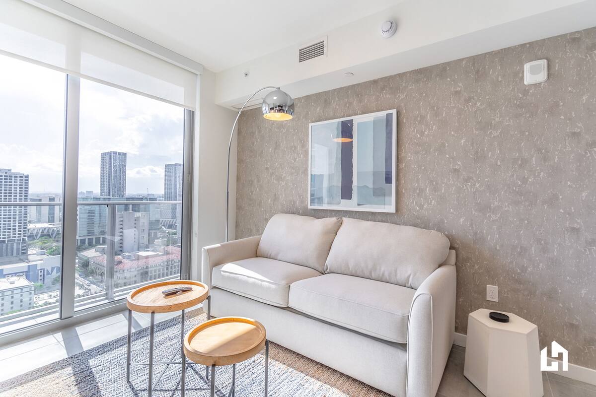 Una sala de estar moderna con un sofá blanco, dos mesitas de madera, una lámpara de pie y vistas a la ciudad a través de amplios ventanales.