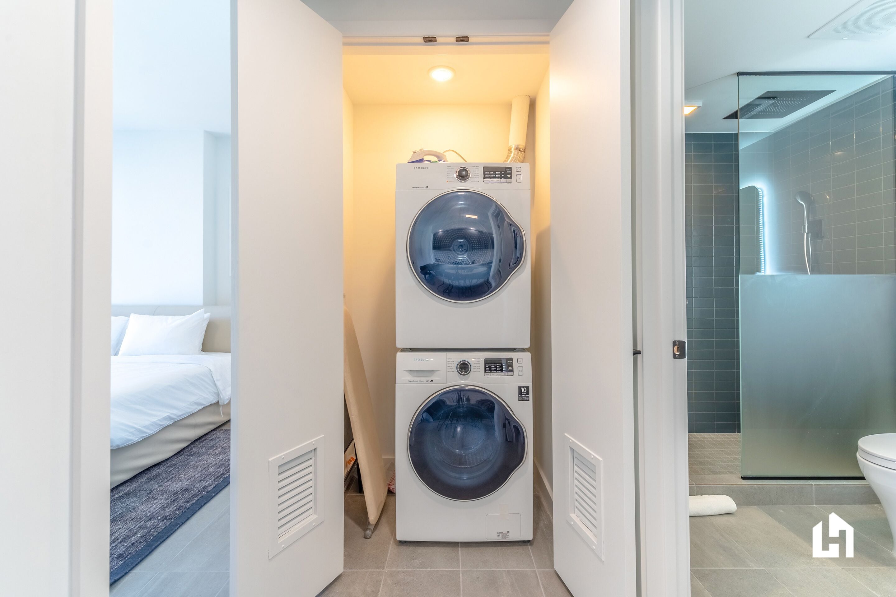 A laundry room with two stacked front-loading washing machines, a bed with white bedding, and a glass shower enclosure.