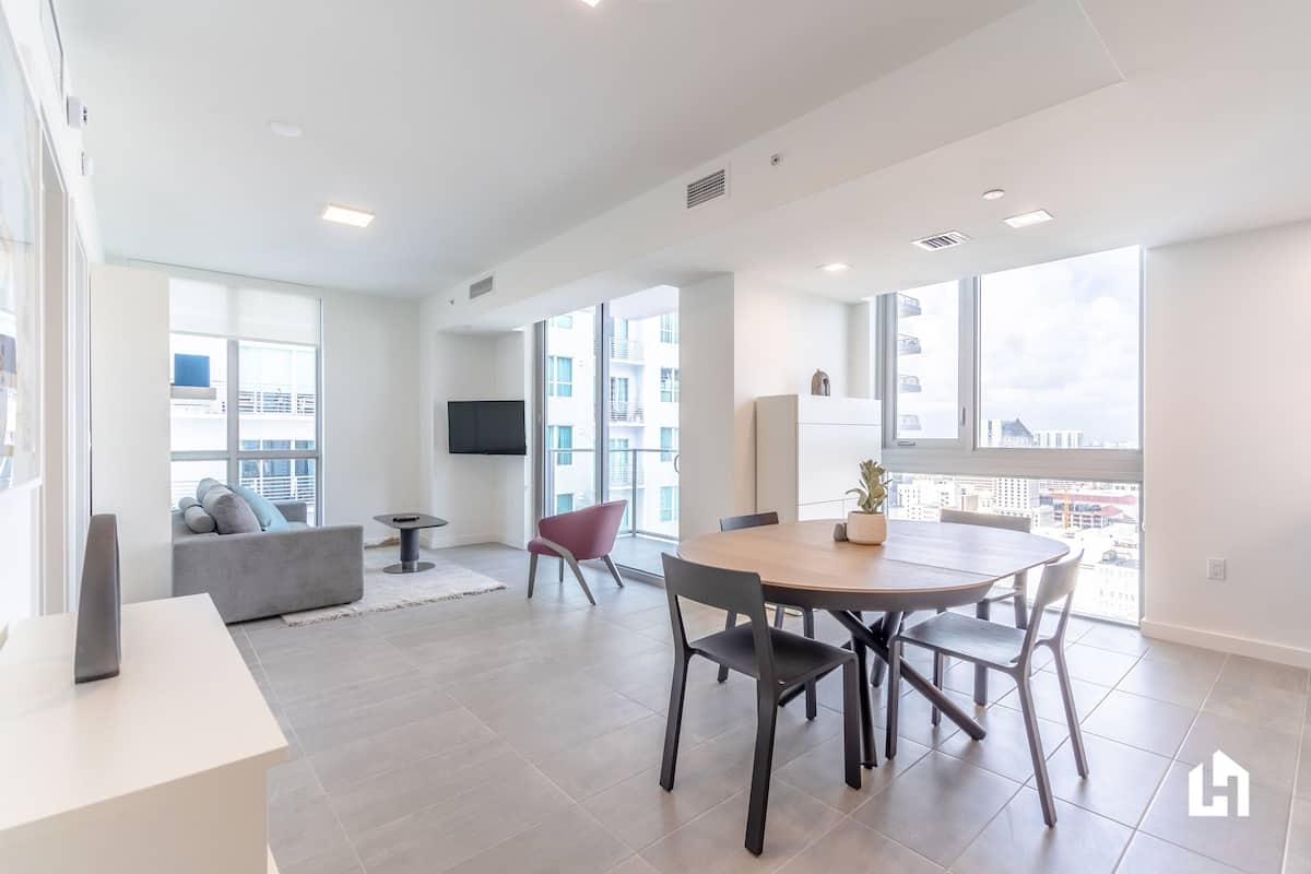 Una sala de estar moderna con un sofá gris, una silla roja, una mesa de comedor de madera con cuatro sillas y ventanas grandes con vistas a la ciudad.