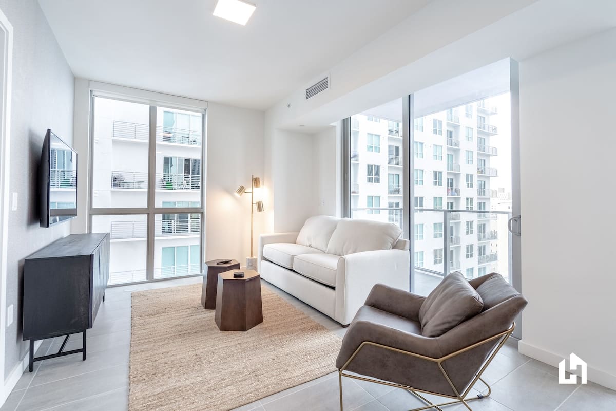 Una sala de estar moderna con un sofá, un sillón, un televisor, ventanas amplias y vistas a un edificio.