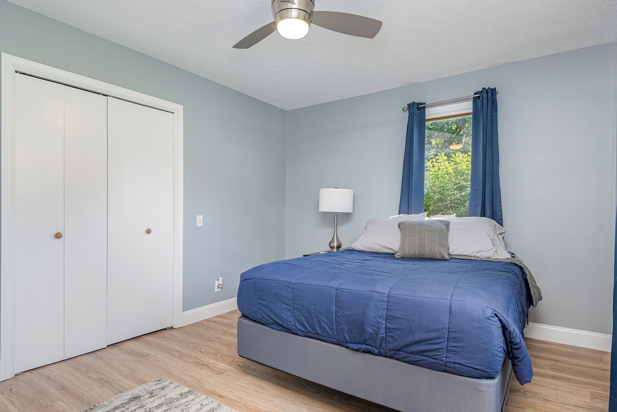 A bedroom with a bed, a ceiling fan, a window with curtains, and a door.
