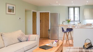 Ein modernes Wohnzimmer mit einer Couch, einem Holztisch und einer Küchenzeile inklusive Kühlschrank, Spüle und einem Fenster mit Jalousien.