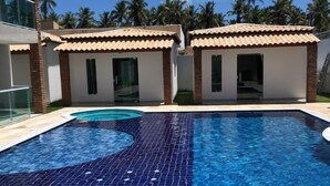A swimming pool with a tiled design, two small buildings with tiled roofs, and palm trees in the background. - Vila Bangalô (São José da Coroa Grande)