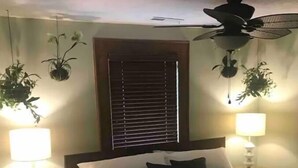 Un dormitorio con cama, dos lámparas de noche, ventilador de techo, ventana con persianas y plantas colgantes.