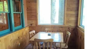 A wooden cabin interior with a dining table and chairs, a window with a view of trees, and a blue-framed window.