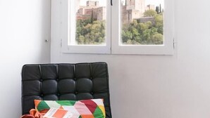 Una stanza con una poltrona in pelle nera, un cuscino colorato e una finestra che si affaccia su edifici e alberi.