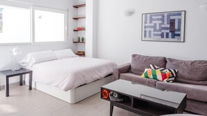 Een moderne slaapkamer met een bed, een grijze bank, een bijzettafeltje, een bureau met lamp, een plafondventilator, een raam en een wandplank met boeken en decoratieve items.