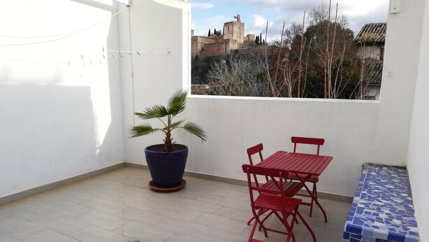 Een klein terras buiten met een betegelde vloer, een potpalm, een rode tafel en stoelen, en een uitzicht op een historisch pand op de achtergrond.