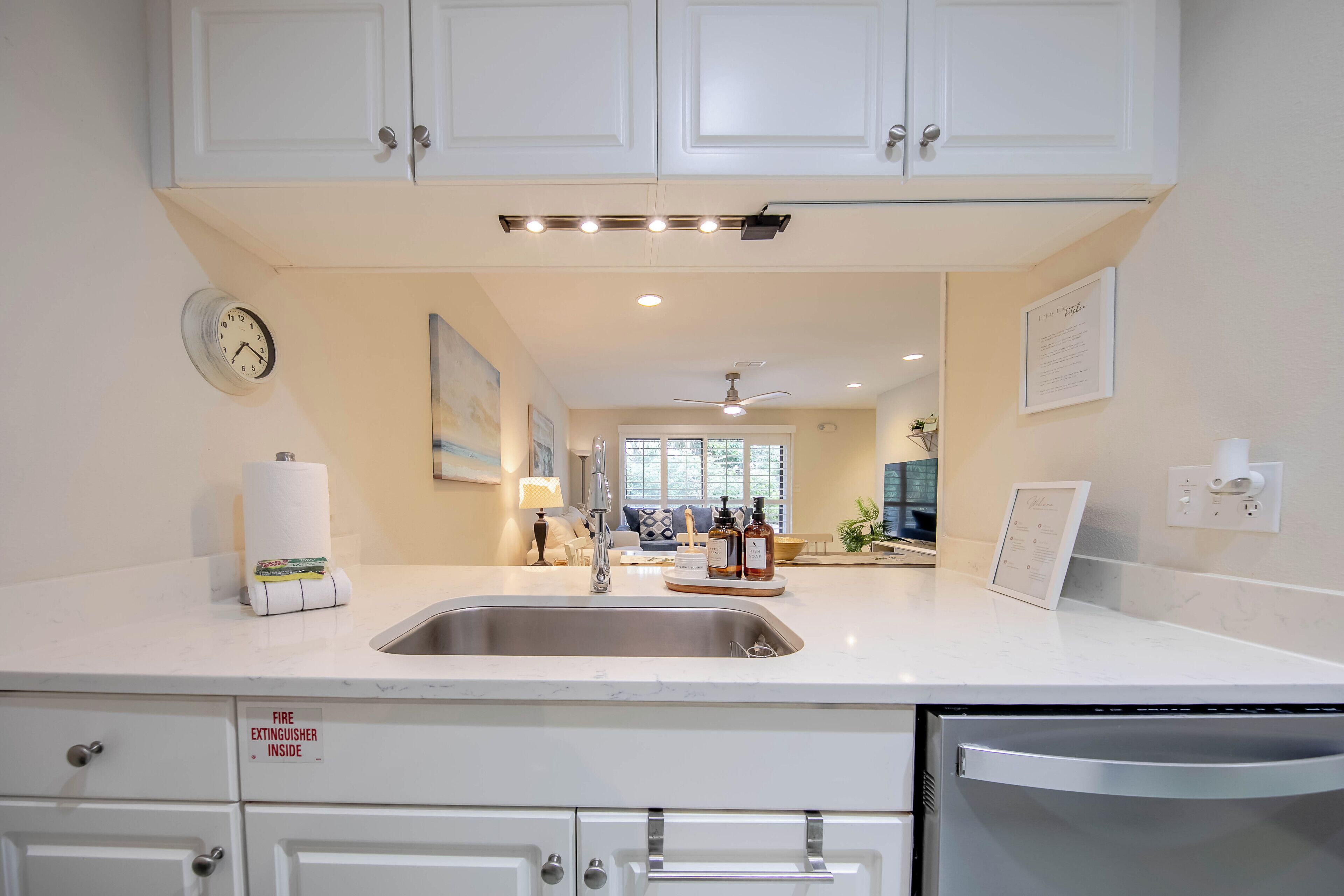 A modern kitchen with white cabinets, a stainless steel sink, and a built-in dishwasher. There is a clock on the wall and a fire extinguisher sign on the counter.