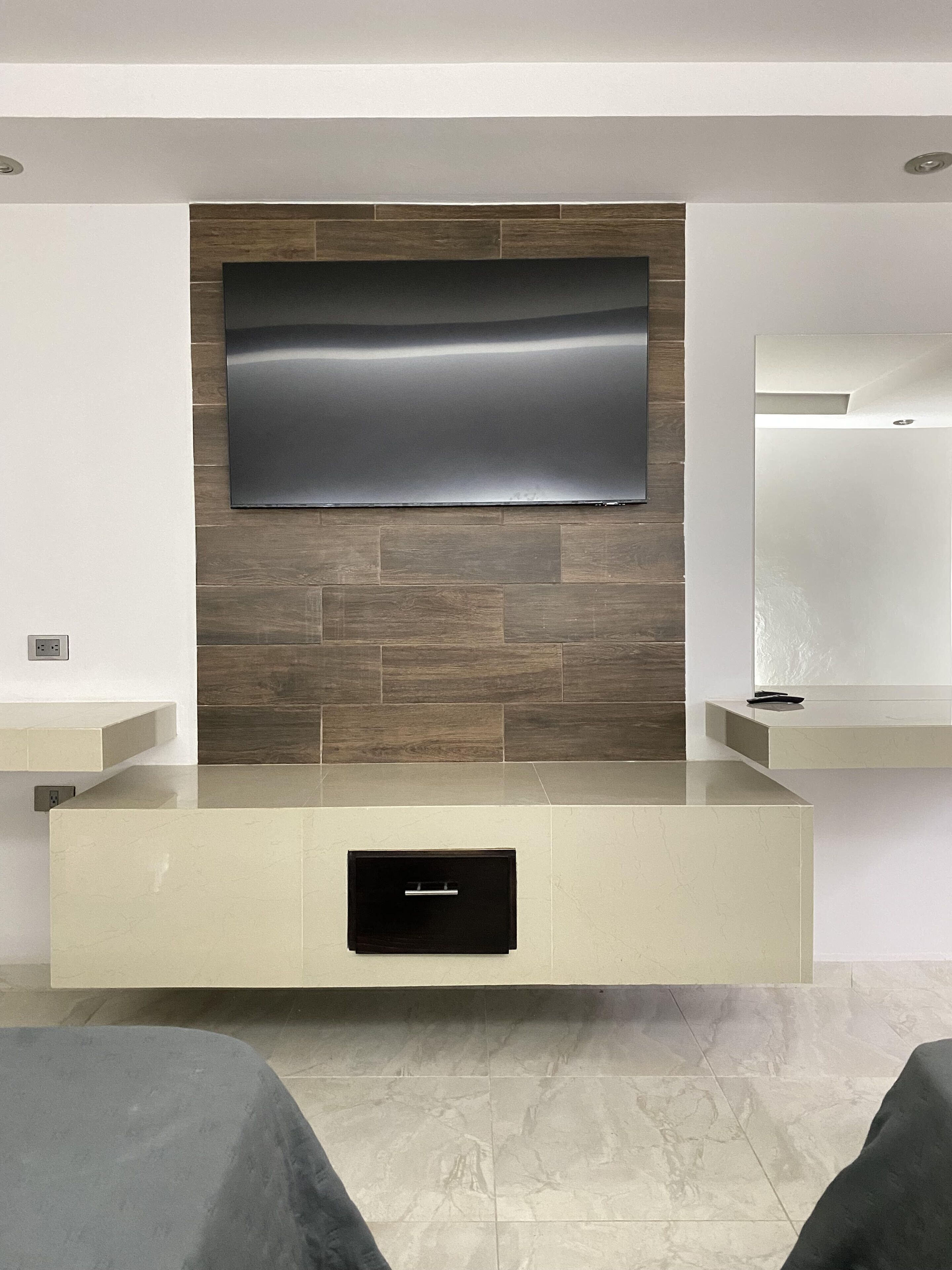 A modern living room with a flat-screen TV mounted on a wooden wall, a built-in bench with a drawer, and a tiled floor.