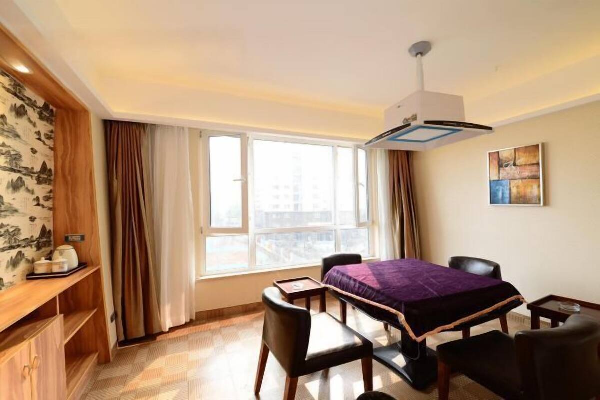 Habitación de hotel con cama, escritorio, silla, mesita, estante con objetos, ventana con cortinas y un cuadro en la pared.