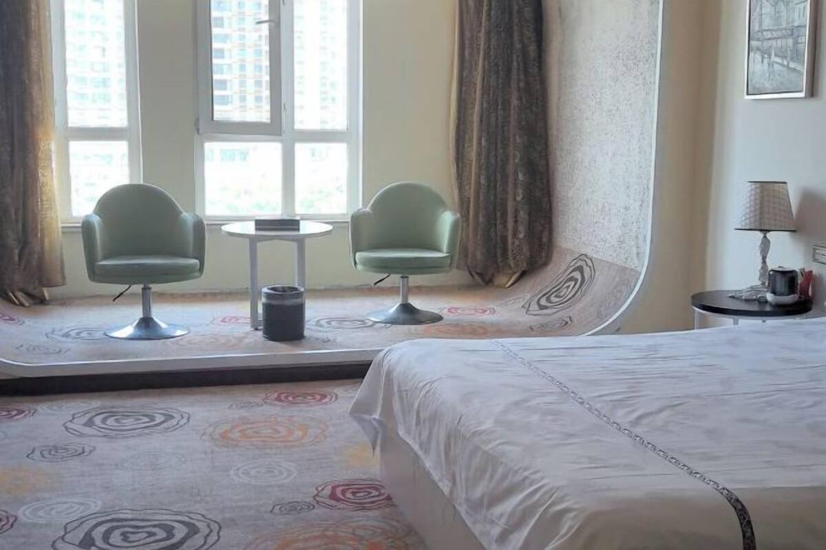 Habitación de hotel con cama, dos sillones, una mesita, lámpara y vistas a edificios a través de amplias ventanas.