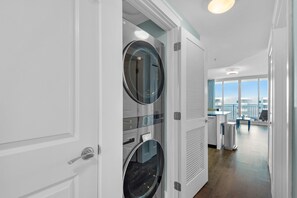 A modern laundry room with two large front-loading washers and dryers, a small table with a vase, and a view of the city through large windows. - Stylish Top-Floor Waterscape Condo, Free Beach Service! (Fort Walton Beach)