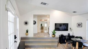 Una sala moderna con un televisor de pantalla plana, una zona de comedor con una mesa y sillas negras, y una escalera que lleva a otra habitación.