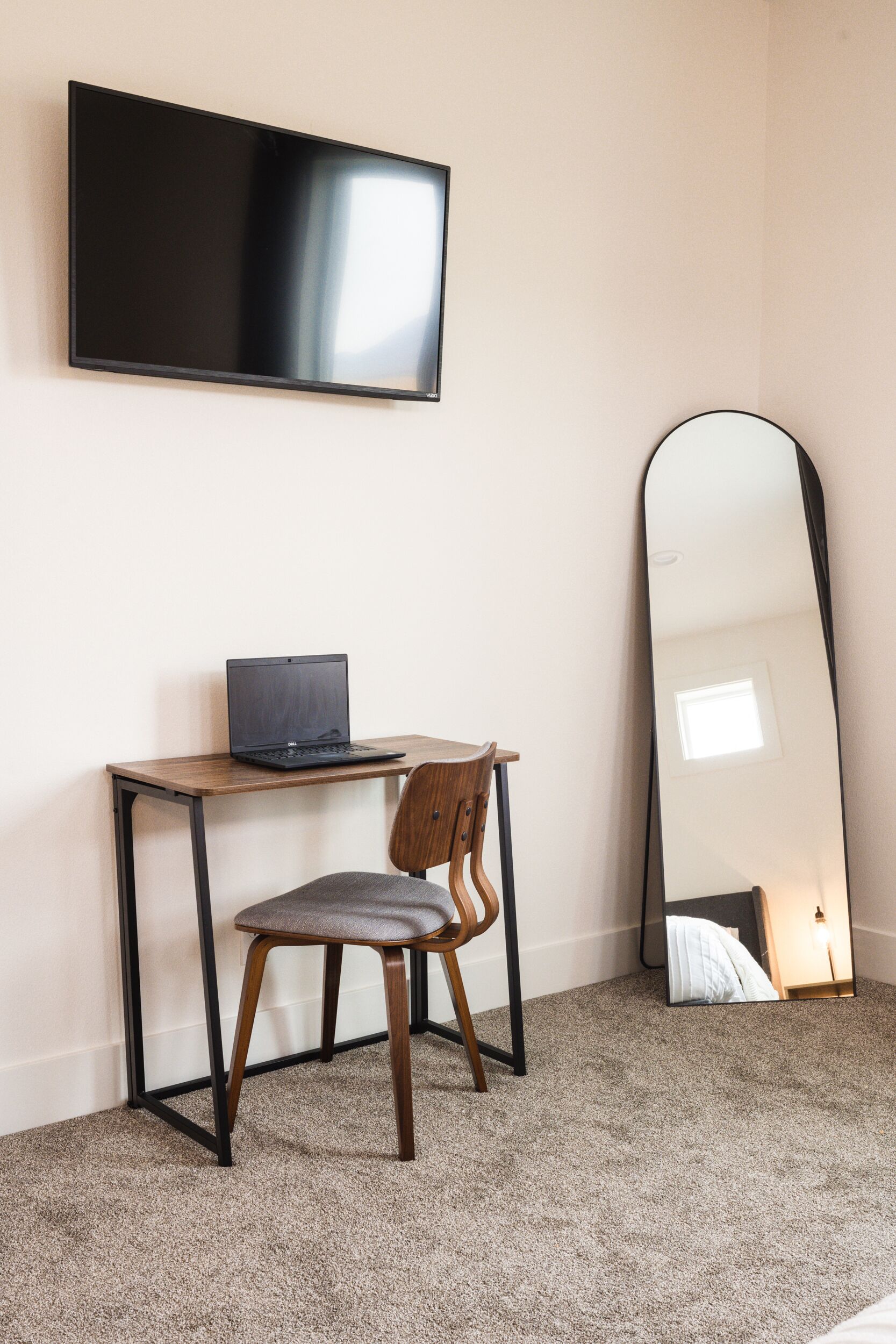 Une pièce dotée d’un téléviseur à écran plat fixé au mur, d’un bureau en bois avec un ordinateur portable, d’une chaise et d’un grand miroir qui reflète la pièce.