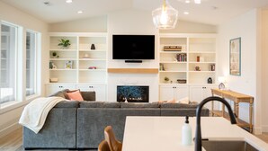 Un salon moderne comprenant un canapé d’angle, une cheminée, des étagères intégrées, un téléviseur à écran plat et un coin repas avec une table et des chaises.
