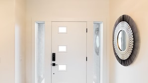 Un hall d’entrée propre et moderne, avec une porte blanche, des panneaux de verre dépoli, un paillasson noir et un miroir décoratif accroché au mur.