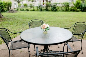 Ein runder Metalltisch mit Glasplatte, umgeben von vier Stühlen aus Mesh-Gewebe, steht auf einer Betonpatio mit Rasenfläche und einem Gebäude im Hintergrund.